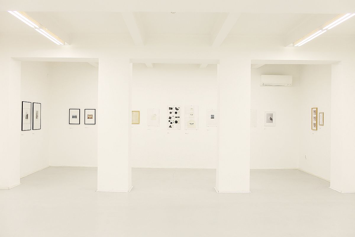 Zdjęcie wnętrza galerii. Ściany, sufit i podłoga w kolorze białym, pośrodku dwie kolumny. Na ścianach zawieszone niewielkie obrazy.